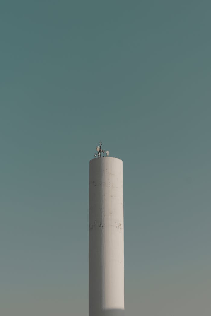 A tall white telecommunications tower stands against a clear blue sky, capturing a minimalist design.